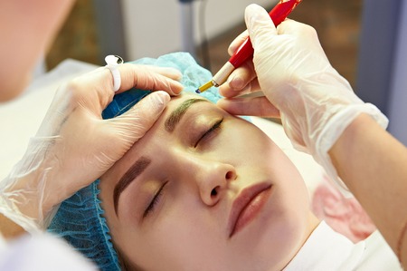 Cosmetician making permanent eyebrow make up for beautiful girl. Eyebrow tattoo. Close up.の写真素材