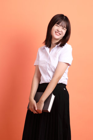 Cheerful female student in Thai university uniform with gesture of holding a textbook isolated on orange background. International Students' Day, World Students' Day. Gen Zの写真素材