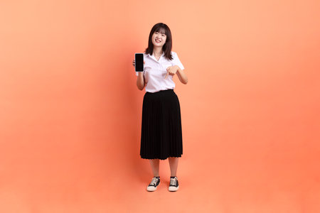 Cheerful female student in Thai university uniform with gesture of Holding mobile phone isolated on orange background. International Students' Day, World Students' Day. Gen Zの写真素材