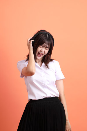 Cheerful female student in Thai university uniform with gesture of wearing headphone isolated on orange background. International Students' Day, World Students' Day. Gen Zの写真素材