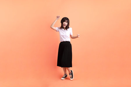 Cheerful female student in Thai university uniform with gesture of wearing headphone isolated on orange background. International Students' Day, World Students' Day. Gen Zの写真素材
