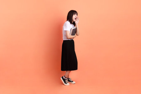 Cheerful female student in Thai university uniform with gesture of holding a textbook isolated on orange background. International Students' Day, World Students' Day.の写真素材