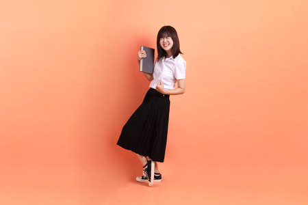 Cheerful female student in Thai university uniform with gesture of holding a textbook isolated on orange background. International Students' Day, World Students' Day.の写真素材