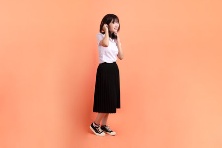 Cheerful female student in Thai university uniform with gesture of wearing headphone isolated on orange background. International Students' Day, World Students' Day. Gen Zの写真素材