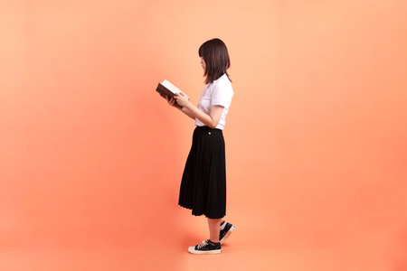 Cheerful female student in Thai university uniform with gesture of holding a textbook isolated on orange background. International Students' Day, World Students' Day. Gen Zの写真素材