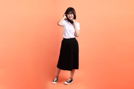 Cheerful female student in Thai university uniform with gesture of wearing headphone isolated on orange background. International Students' Day, World Students' Day. Gen Zの写真素材