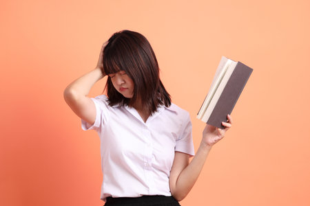 Cheerful female student in Thai university uniform with gesture of holding a textbook isolated on orange background. International Students' Day, World Students' Day. Gen Zの写真素材
