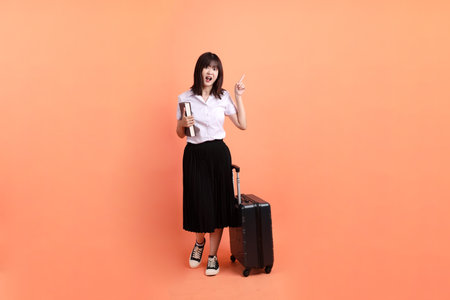 Cheerful female student in Thai university uniform with gesture of holding suitcase isolated on orange background. International Students' Day, World Students' Day.の写真素材