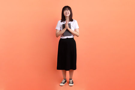 Cheerful female student in Thai university uniform with gesture of holding a textbook isolated on orange background. International Students' Day, World Students' Day. Gen Zの写真素材