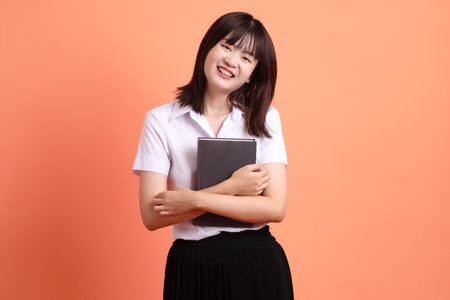 Cheerful female student in Thai university uniform with gesture of holding a textbook isolated on orange background. International Students' Day, World Students' Day. Gen Zの写真素材