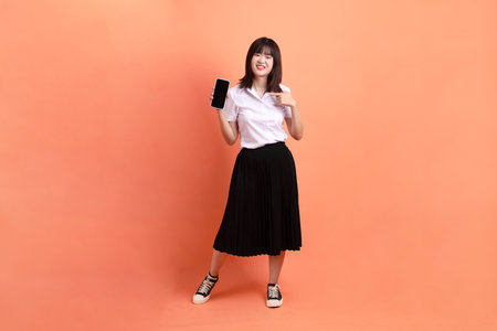 Cheerful female student in Thai university uniform with gesture of Holding mobile phone isolated on orange background. International Students' Day, World Students' Day. Gen Zの写真素材