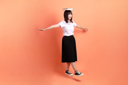 Cheerful female student in Thai university uniform with gesture of holding a textbook isolated on orange background. International Students' Day, World Students' Day. Gen Zの写真素材