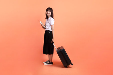 Cheerful female student in Thai university uniform with gesture of holding suitcase isolated on orange background. International Students' Day, World Students' Day.の写真素材