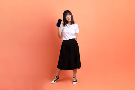 Cheerful female student in Thai university uniform with gesture of Holding mobile phone isolated on orange background. International Students' Day, World Students' Day. Gen Zの写真素材