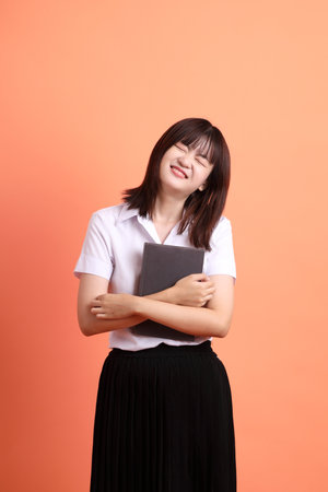 Cheerful female student in Thai university uniform with gesture of holding a textbook isolated on orange background. International Students' Day, World Students' Day. Gen Zの写真素材