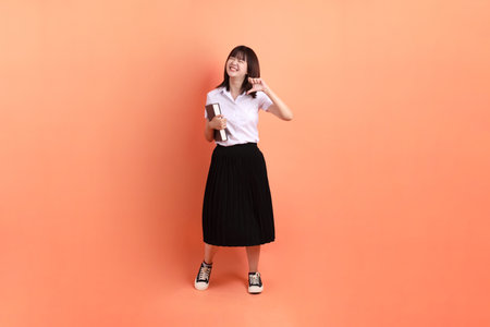 Cheerful female student in Thai university uniform with gesture of holding a textbook isolated on orange background. International Students' Day, World Students' Day.の写真素材