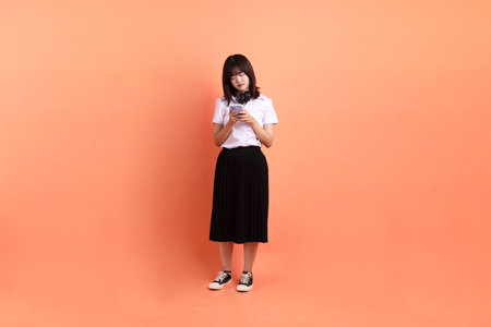 Cheerful female student in Thai university uniform with gesture of holding mobile phone and headphone isolated on orange background. International Students' Day, World Students' Day. Gen Zの写真素材