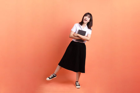 Cheerful female student in Thai university uniform with gesture of holding a textbook isolated on orange background. International Students' Day, World Students' Day. Gen Zの写真素材
