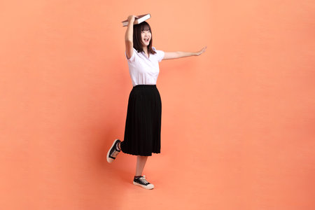 Cheerful female student in Thai university uniform with gesture of holding a textbook isolated on orange background. International Students' Day, World Students' Day. Gen Zの写真素材