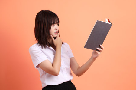 Cheerful female student in Thai university uniform with gesture of holding a textbook isolated on orange background. International Students' Day, World Students' Day. Gen Zの写真素材