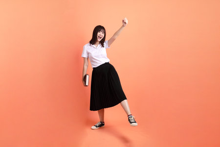 Cheerful female student in Thai university uniform with gesture of holding a textbook isolated on orange background. International Students' Day, World Students' Day. Gen Zの写真素材