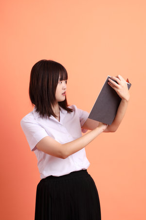 Cheerful female student in Thai university uniform with gesture of holding a textbook isolated on orange background. International Students' Day, World Students' Day. Gen Zの写真素材