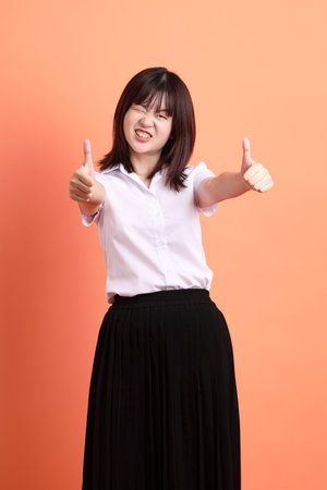 Cheerful female student in Thai university uniform with gesture of thumbs up isolated on orange background. International Students' Day, World Students' Day.の写真素材