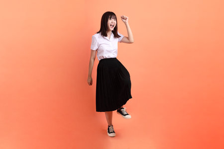 Cheerful female student in Thai university uniform with gesture of walking isolated on orange background. International Students' Day, World Students' Day. Gen Zの写真素材