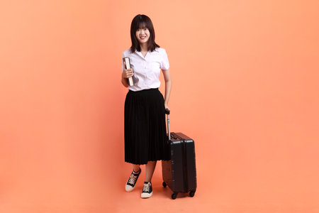 Cheerful female student in Thai university uniform with gesture of holding suitcase isolated on orange background. International Students' Day, World Students' Day.の写真素材