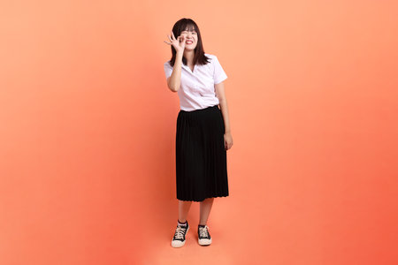 Cheerful female student in Thai university uniform with gesture of ok or confirm isolated on orange background. International Students' Day, World Students' Day. Gen Zの写真素材