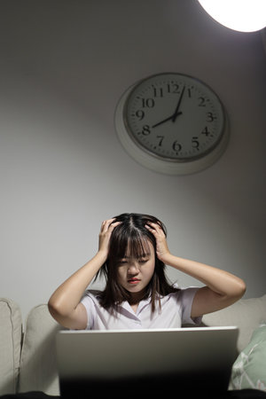 A young female student in a Thai university uniform sits on a sofa in the living room, studying online with a laptop. International Students' Day, World Students' Day. Gen Z, E-learningの写真素材