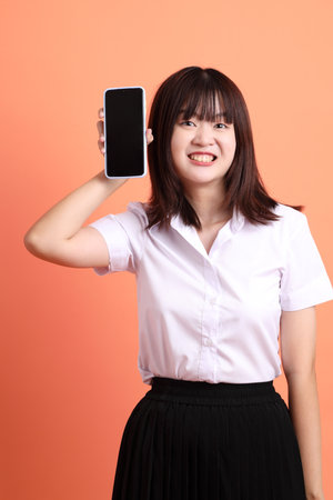 Cheerful female student in Thai university uniform with gesture of Holding mobile phone isolated on orange background. International Students' Day, World Students' Day. Gen Zの写真素材