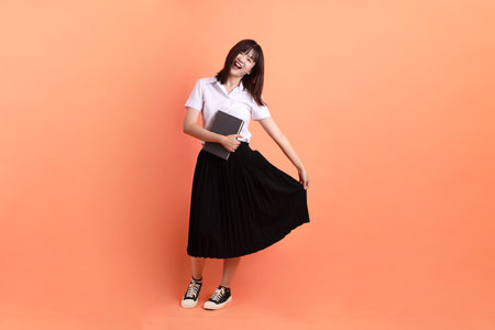 Cheerful female student in Thai university uniform with gesture of holding a textbook isolated on orange background. International Students' Day, World Students' Day. Gen Zの写真素材