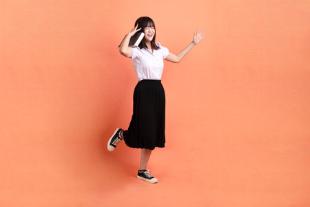 Cheerful female student in Thai university uniform with gesture of holding a textbook isolated on orange background. International Students' Day, World Students' Day. Gen Zの写真素材
