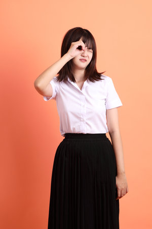 Cheerful female student in Thai university uniform with gesture of looking far away isolated on orange background. International Students' Day, World Students' Day. Gen Zの写真素材