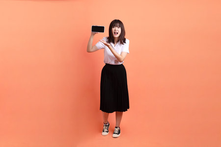Cheerful female student in Thai university uniform with gesture of Holding mobile phone isolated on orange background. International Students' Day, World Students' Day. Gen Zの写真素材