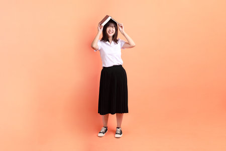 Cheerful female student in Thai university uniform with gesture of holding a textbook isolated on orange background. International Students' Day, World Students' Day. Gen Zの写真素材