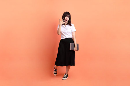 Cheerful female student in Thai university uniform with gesture of holding a textbook isolated on orange background. International Students' Day, World Students' Day. Gen Zの写真素材