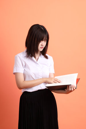 Cheerful female student in Thai university uniform with gesture of holding a textbook isolated on orange background. International Students' Day, World Students' Day. Gen Zの写真素材