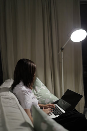 A young female student in a Thai university uniform sits on a sofa in the living room, studying online with a laptop. International Students' Day, World Students' Day. Gen Z, E-learningの写真素材