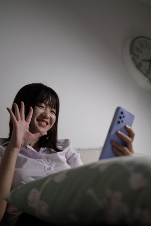 A young female student in a Thai university uniform sits on a sofa in the living room, studying online with an application on mobile phone. International Students' Day, Gen Z, E-learningの写真素材