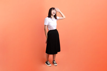 Cheerful female student in Thai university uniform with gesture of looking far away isolated on orange background. International Students' Day, World Students' Day. Gen Zの写真素材