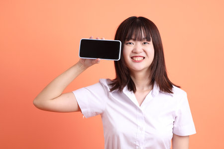 Cheerful female student in Thai university uniform with gesture of Holding mobile phone isolated on orange background. International Students' Day, World Students' Day. Gen Zの写真素材