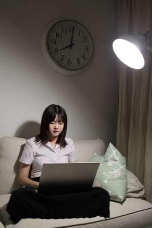A young female student in a Thai university uniform sits on a sofa in the living room, studying online with a laptop. International Students' Day, World Students' Day. Gen Z, E-learningの写真素材