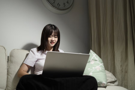 A young female student in a Thai university uniform sits on a sofa in the living room, studying online with a laptop. International Students' Day, World Students' Day. Gen Z, E-learningの写真素材