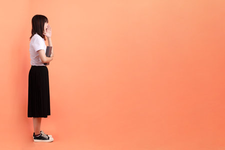 Cheerful female student in Thai university uniform with gesture of holding a textbook isolated on orange background. International Students' Day, World Students' Day. Gen Zの写真素材
