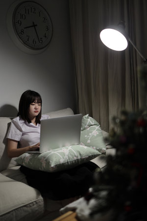 A young female student in a Thai university uniform sits on a sofa in the living room, studying online with a laptop. International Students' Day, World Students' Day. Gen Z, E-learningの写真素材