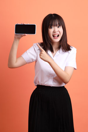 Cheerful female student in Thai university uniform with gesture of Holding mobile phone isolated on orange background. International Students' Day, World Students' Day. Gen Zの写真素材
