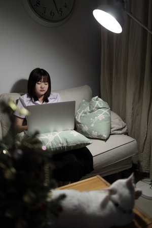 A young female student in a Thai university uniform sits on a sofa in the living room, studying online with a laptop. International Students' Day, World Students' Day. Gen Z, E-learningの写真素材