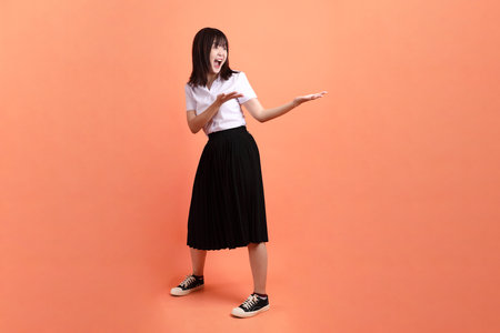 Cheerful female student in Thai university uniform with gesture of pointing isolated on orange background. International Students' Day, World Students' Day. Gen Zの写真素材